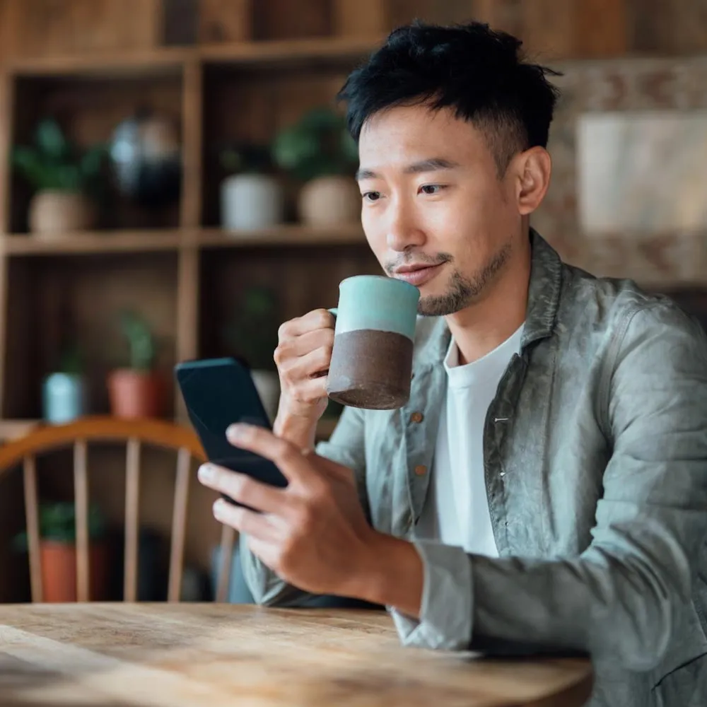 A man sitting at the table drinking out of a coffee mug and using his phone 