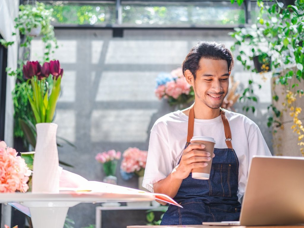 A man surrounded by flowers with a coffee cup, smiling and looking at his laptop