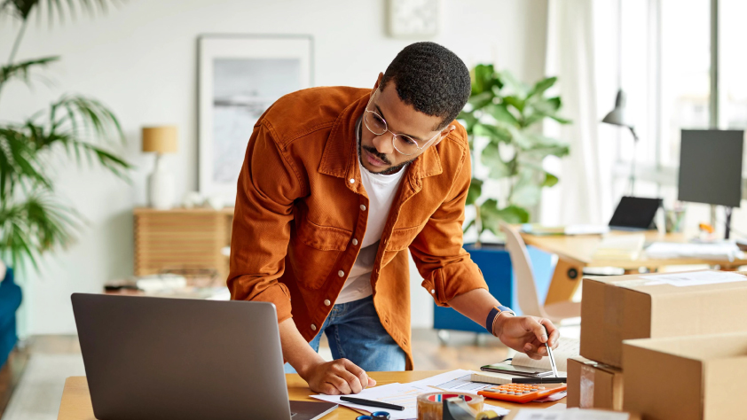 Personne debout à un bureau à la maison, lisant sur son ordinateur portable et prenant des notes