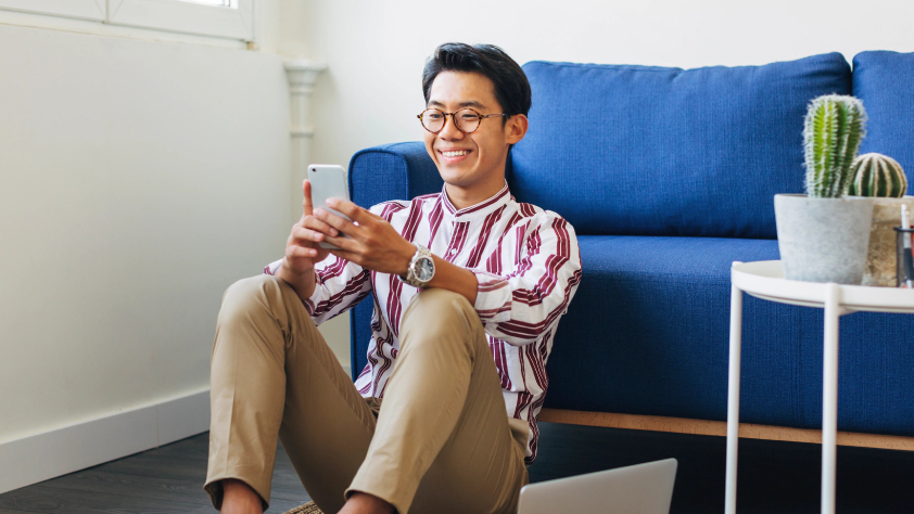 Personne souriante assise par terre, le dos appuyé contre un canapé, en train de consulter des messages sur WhatsApp