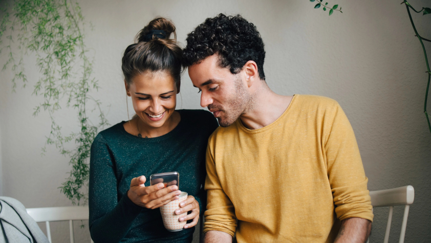 Deux personnes assises à l’intérieur sur un banc. L’une tient un café dans la main et elles regardent toutes les deux un message professionnel sur WhatsApp.