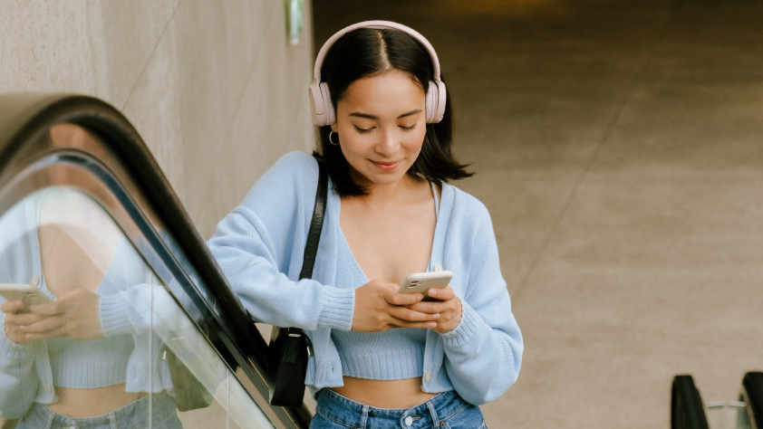 Person riding up escalator wearing a blue sweater, jeans, and pink headphones. They are carrying a black purse and reading a business message on WhatsApp on phone.