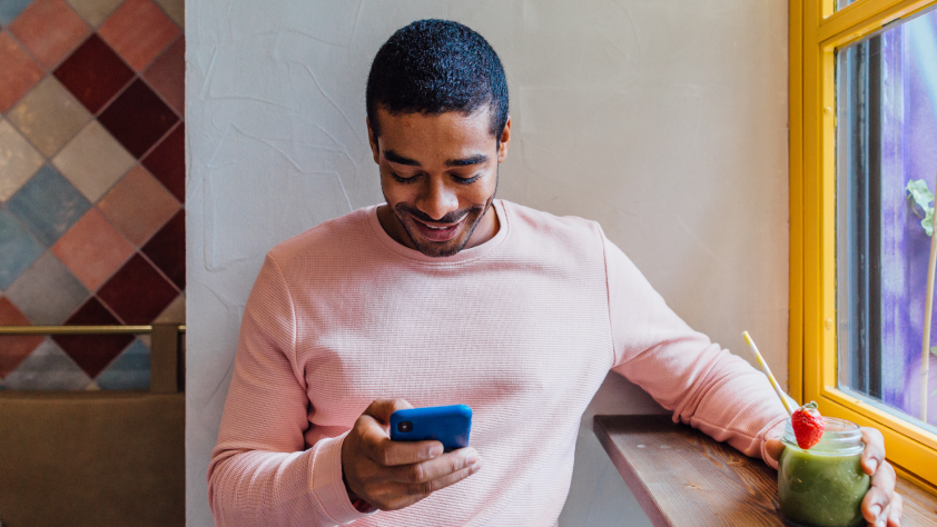 Personne assise dans un café devant la fenêtre, portant un t-shirt rose à manches longues. Elle tient un matcha latte et sourit en regardant son téléphone.