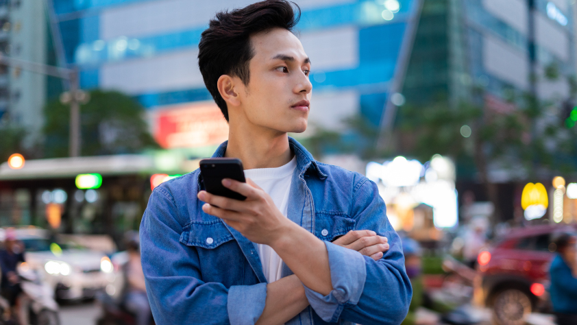 Personne debout devant un arrière-plan représentant une ville animée. Elle porte un t-shirt blanc et une chemise en jean bleue, tient un téléphone et regarde au loin.