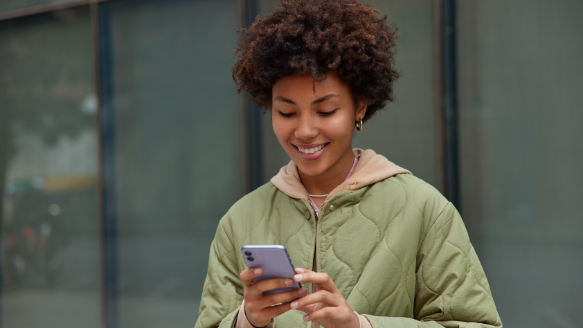 Personne debout devant les fenêtres d’un bâtiment de bureaux. Elle porte une veste vert clair et sourit en regardant un iPhone violet.