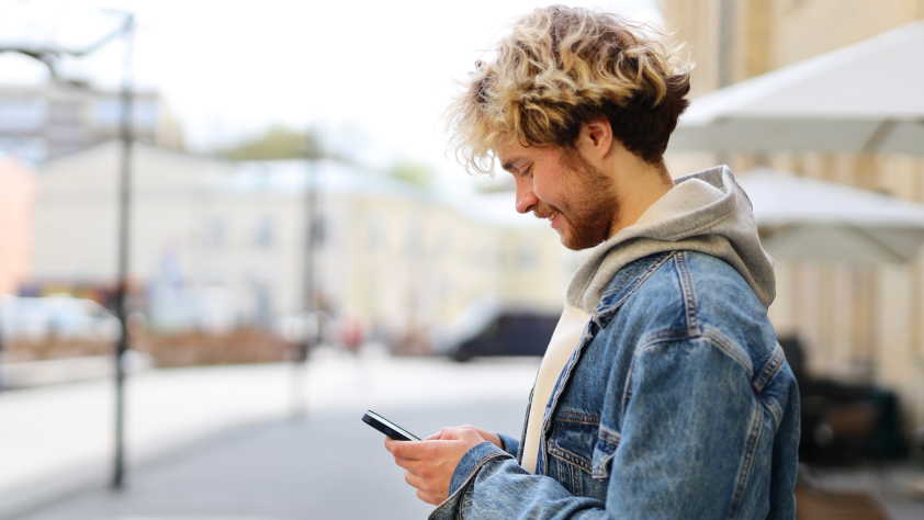 Personne debout en dehors d’un café avec des parapluies, un bâtiment et une rue en arrière-plan. Ils portent un sweat-shirt et une veste en jean tout en regardant une recommandation de produit via WhatsApp sur téléphone.