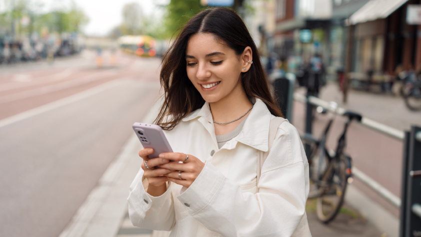 Personne debout sur la rue Amsterdam avec la ville en arrière-plan. Elle porte une veste blanche tout en souriant et en regardant son smartphone.