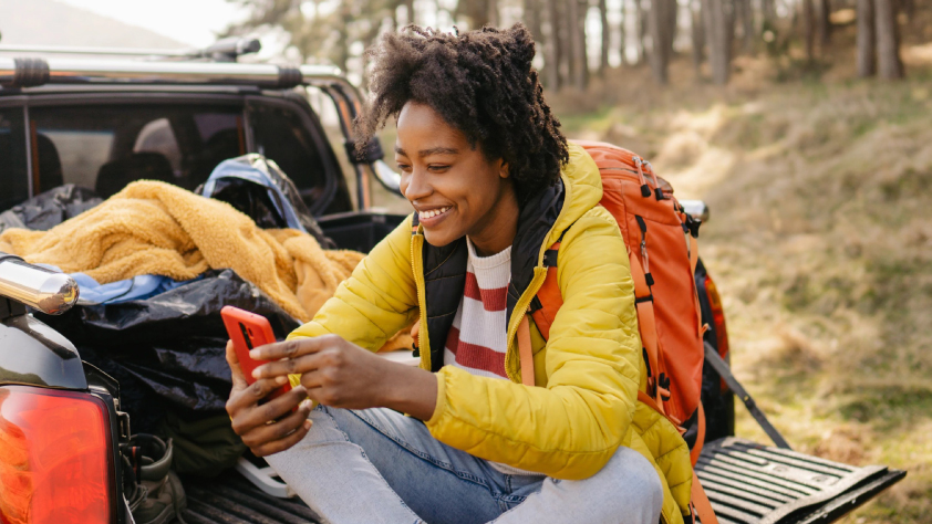 Personne souriante assise à l’arrière d’un pick-up avec du matériel de camping dans les bois, pendant un appel vidéo