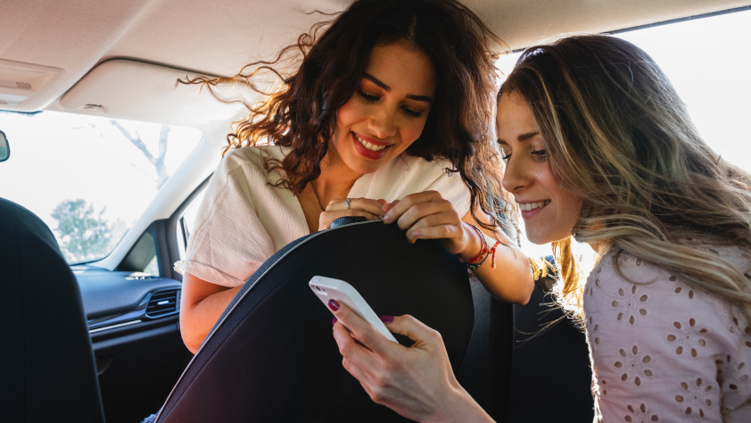 Deux personnes dans une voiture. Une personne tournée vers l’avant depuis la place passager regarde le téléphone de la personne assise à l’arrière