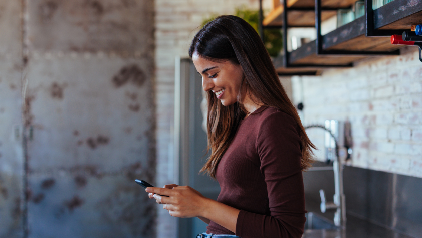 Une personne debout dans une cuisine, le bras appuyé sur le comptoir, qui sourit en lisant un message professionnel sur WhatsApp.