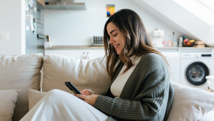 Personne assise sur un canapé blanc, portant un t-shirt blanc et un pull vert. Elle sourit en regardant une publicité sur Instagram sur son téléphone.