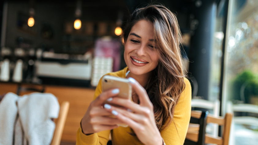 Persona sentada en un café con una camisa amarilla. Sonríe y habla con un equipo de soporte por WhatsApp en un iPhone.