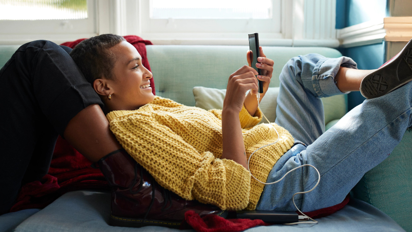 Person laying on couch leaning on someone's legs while smiling at phone 