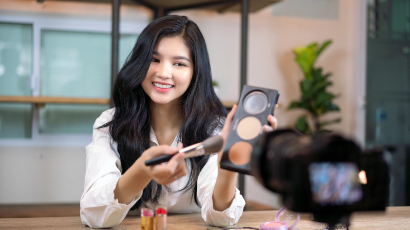 Person smiling and showing a makeup palette and brush while filming a makeup tutorial 