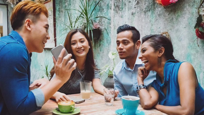 Empat orang duduk mengelilingi meja di kafe sementara salah satu menunjukkan sesuatu di teleponnya
