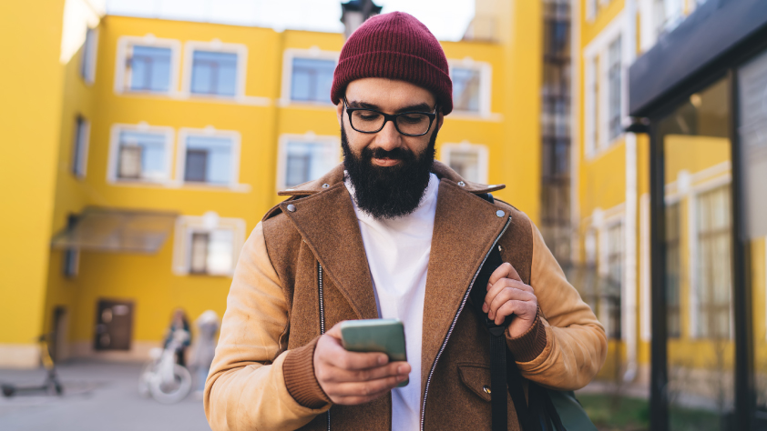 Persona caminando por la calle con edificios amarillos de fondo. Lleva un gorro bordó, lentes y una chaqueta marrón mientras revisa un catálogo de productos en WhatsApp.