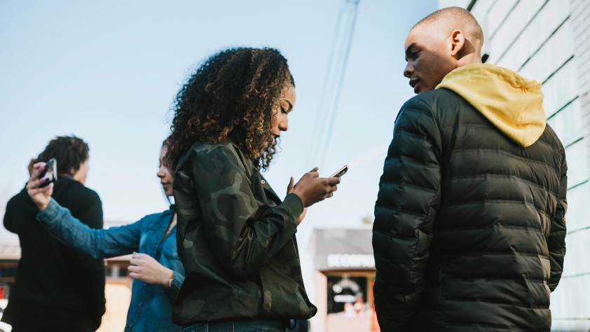 Une personne prend une photo sur un téléphone en arrière-plan tandis que deux personnes sont debout devant et que l’une fait défiler le téléphone