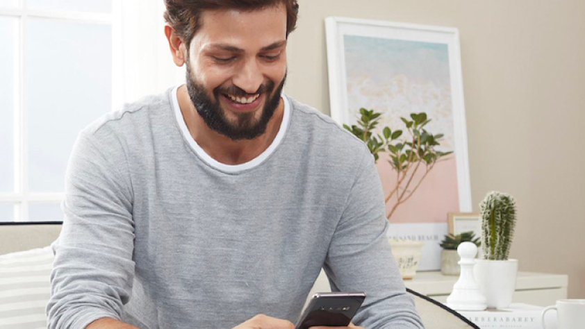 Person sitting in living room with white furniture. They are wearing a gray long sleeve shirt and smiling while reading a personalized WhatsApp message from Tata CLiQ.