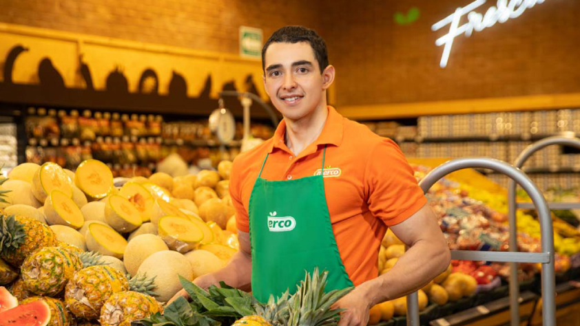 Employé d’un supermarché Merco souriant, debout devant la section des fruits