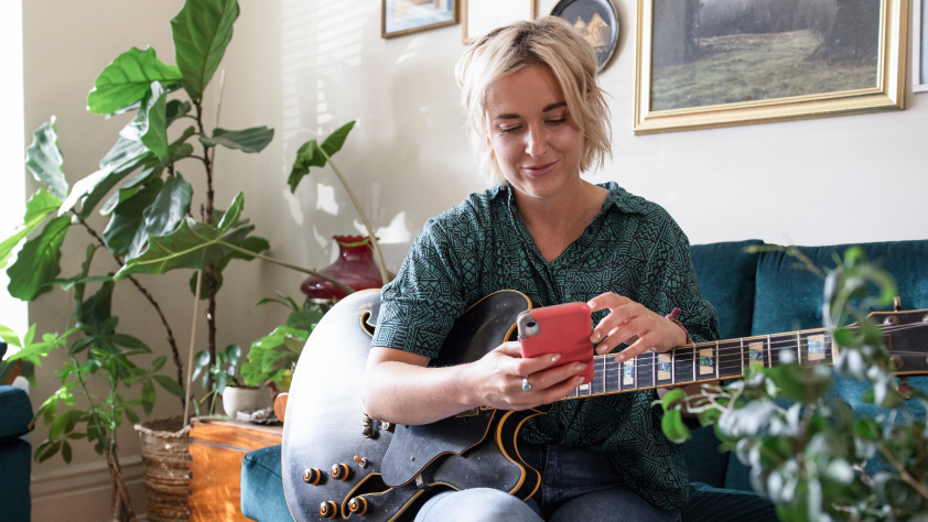 Person sitting on a blue couch surrounded by plants in the living room while holding a guitar and reading a business survey sent through WhatsApp 