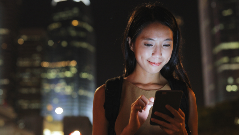 Une personne marchant dans une ville la nuit tout en regardant son téléphone. La lumière du téléphone éclaire son visage.