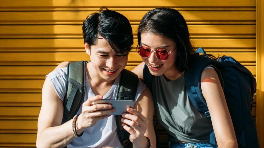 Dua orang mengenakan ransel tersenyum sambil menonton iklan di telepon