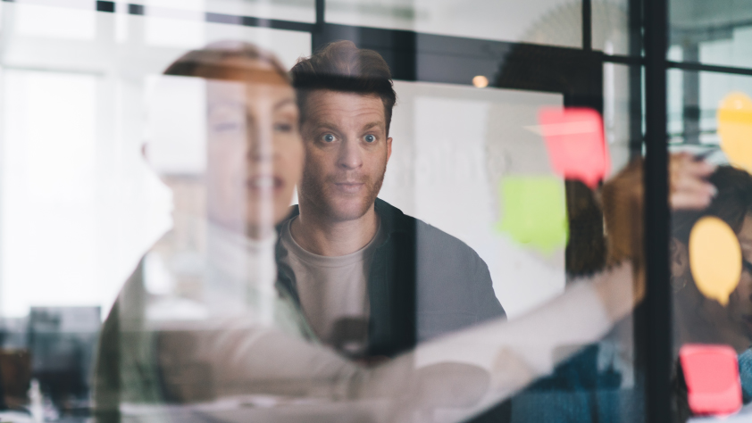 Quelques personnes dans une salle de conférence lors d’une session de brainstorming. L’une des personnes écrit une idée sur la vitre tandis que l’autre la regarde avec les yeux grands ouverts.