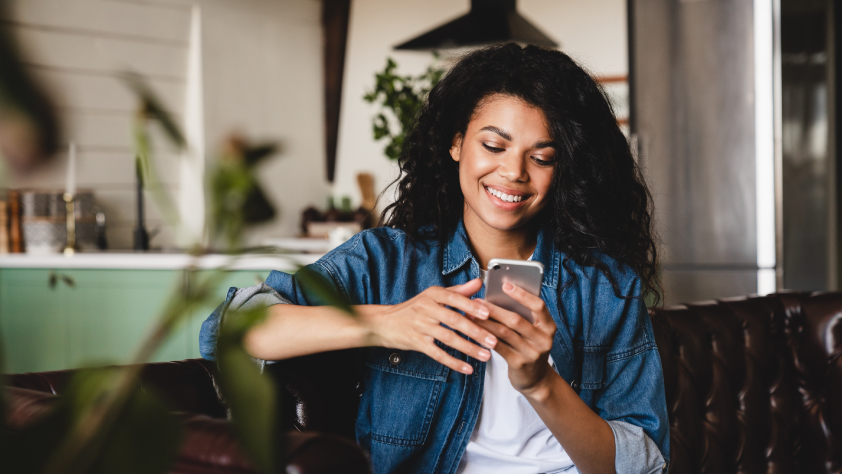 Personne assise sur un canapé marron, portant un t-shirt blanc et une chemise en jean bleue. Elle interagit avec une publicité clic vers WhatsApp sur son téléphone.