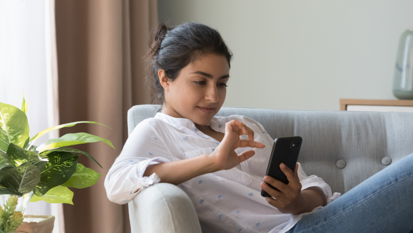 Personne assise dans le salon sur un canapé bleu clair, portant une chemise blanche et un jean bleu. Ils interagissent avec une publicité qui clique vers WhatsApp.