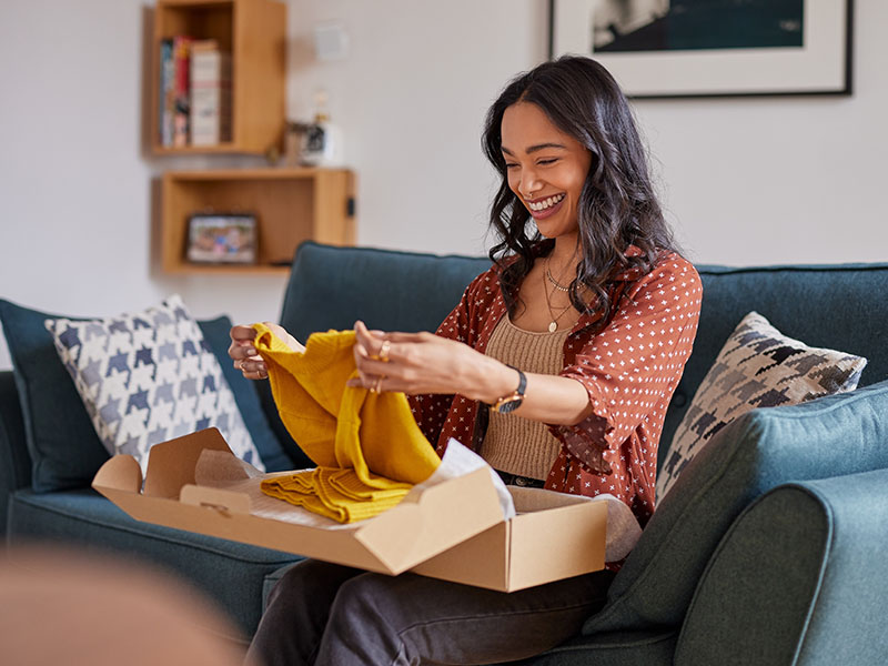 Une femme souriante assise sur un canapé, déballant un cadeau pour révéler un pull jaune.