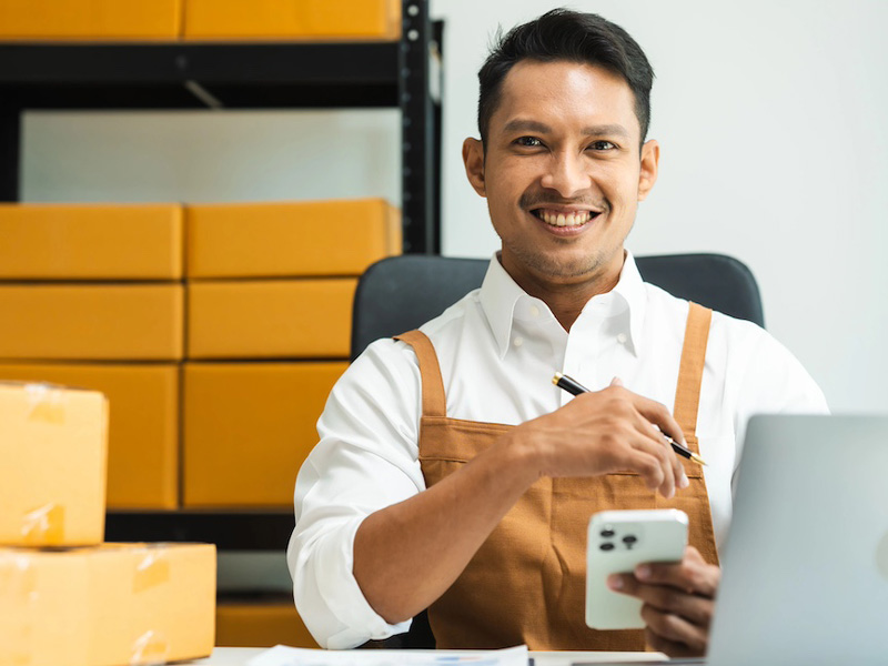 Business owner smiling at the camera, working from his phone and laptop