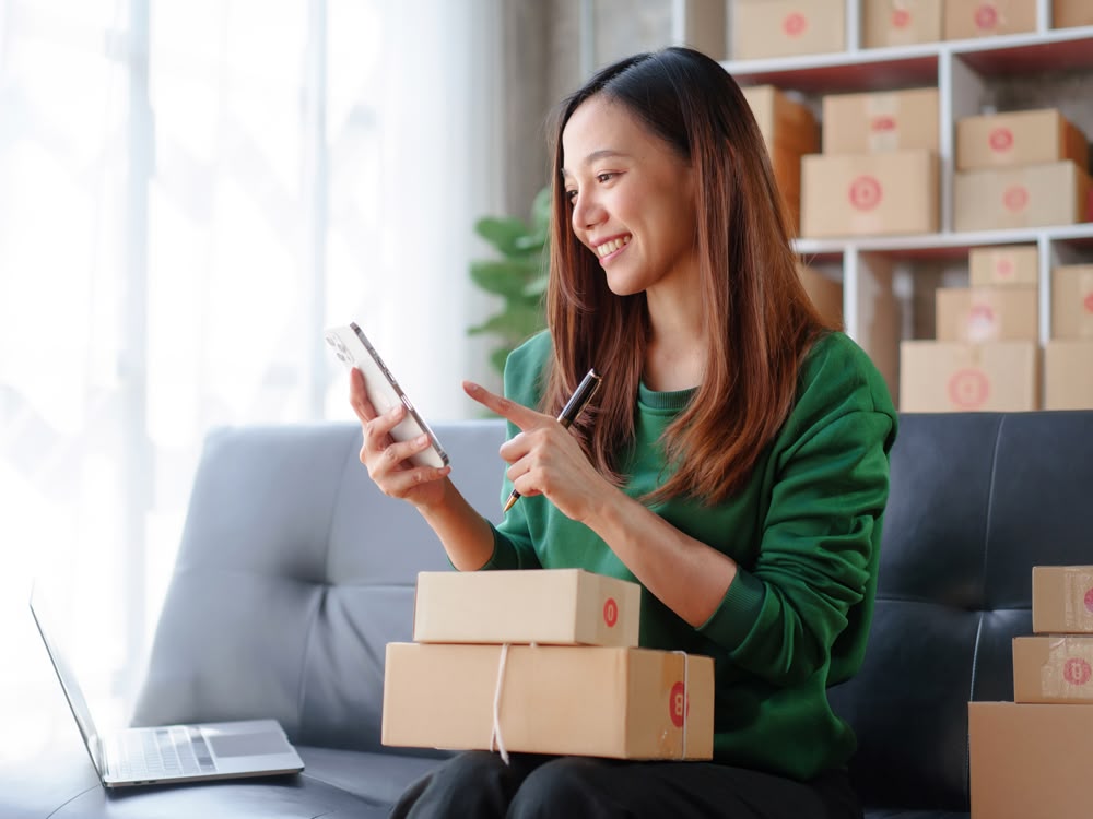 A woman sitting on a chair with packages in her lap, holding a phone and smiling
