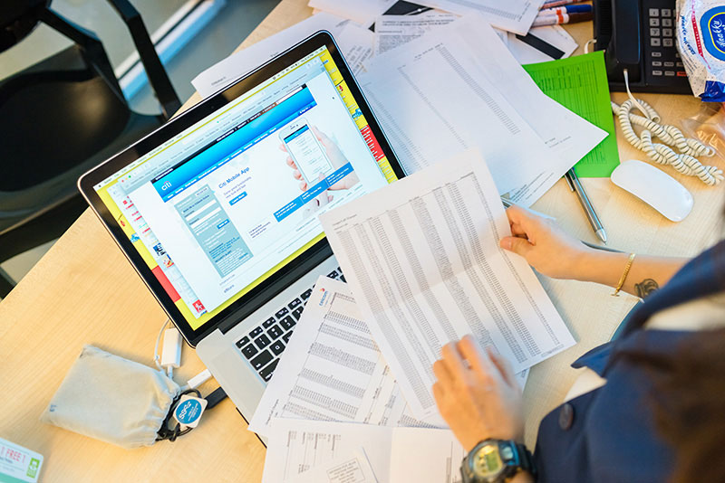 A person sitting at a desk, with their back to the camera, working on a laptop surrounded by scattered papers
