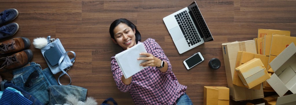 Pemandangan dari atas seseorang yang berbaring di lantai sambil mencatat dan dikelilingi laptop, ponsel, kotak, dan pakaian