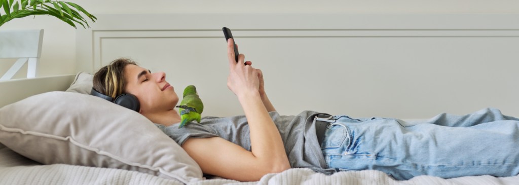 Seseorang berbaring di tempat tidur mengenakan headphone dan menggulir ponsel dengan seekor burung hijau berdiri di pundaknya