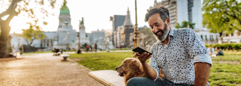 Orang duduk bersama anjing di pusat kota sambil tersenyum dan melihat telepon