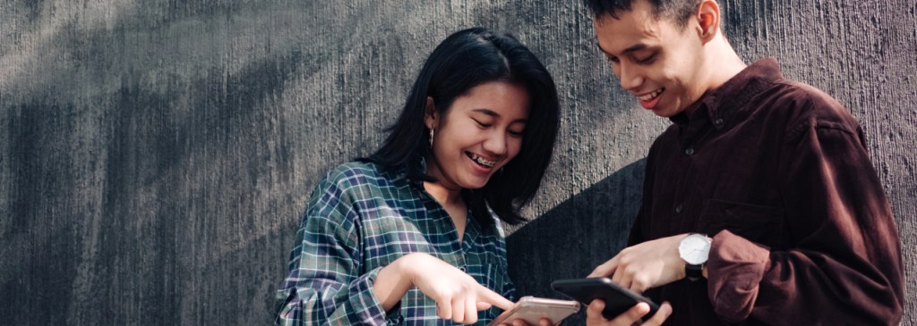 Dua orang berdiri di depan dinding sambil menunjukkan iklan dengan menunjuk telepon mereka