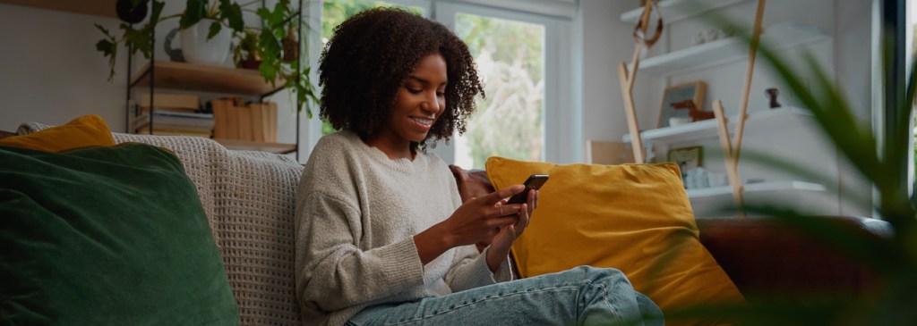 Seseorang duduk di sofa dengan bantal hijau dan kuning di ruang tamu sambil tersenyum dan membaca pesan bisnis baru via WhatsApp