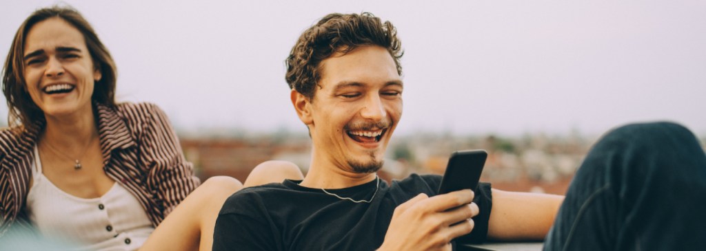 Dua orang duduk di luar sambil tertawa, sementara orang di depan sedang menggulir teleponnya