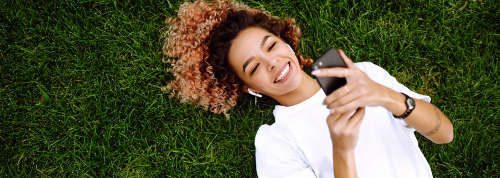 Une personne allongée dans l’herbe portant des AirPods et un t-shirt blanc. Elle sourit et clique sur une publicité qui la redirige vers WhatsApp sur son téléphone.