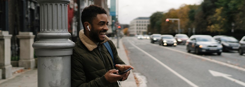 Persona con una chaqueta verde oscuro apoyada en un farol en la calle. Están riendo, llevan puestos los AirPods y sostienen el teléfono.