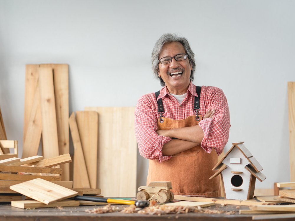 Man smiling in a wood shop next to a wooden bird house and miniature monster truck