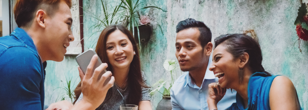 Empat orang duduk mengelilingi meja makan. Seseorang menunjukkan pesan WhatsApp di layar teleponnya kepada teman-temannya.