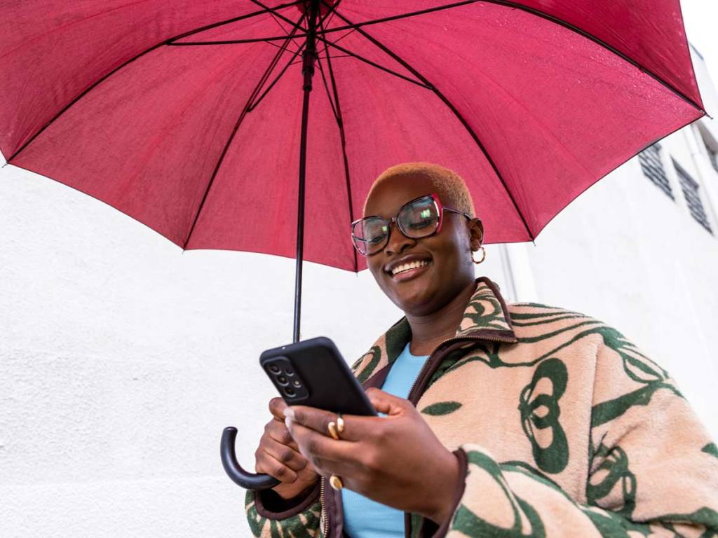 Une femme tenant un parapluie rouge qui marche dehors et qui sourit en regardant son téléphone