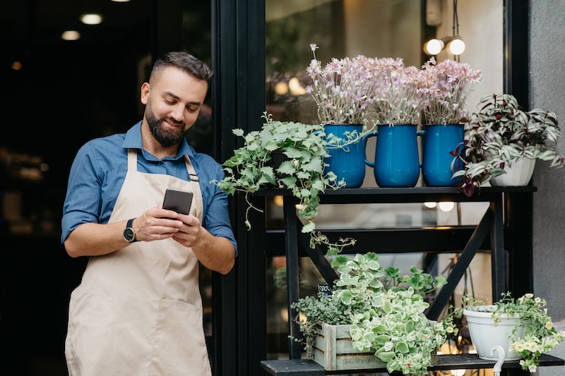 Portrait of young caucasian male owner of flower center typing on phone, employee manager in apron doing inventory. Cheerful man use smartphone near flower shop with plants in pots and vases outdoor.