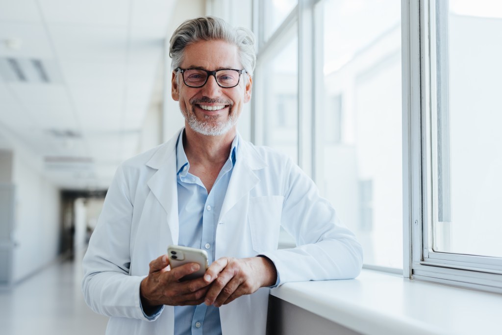 Un médecin d’âge mûr, aux cheveux gris et portant des lunettes, se tient dans une clinique médicale, souriant, en blouse blanche avec un stéthoscope. Il regarde la caméra tout en tenant son téléphone mobile.