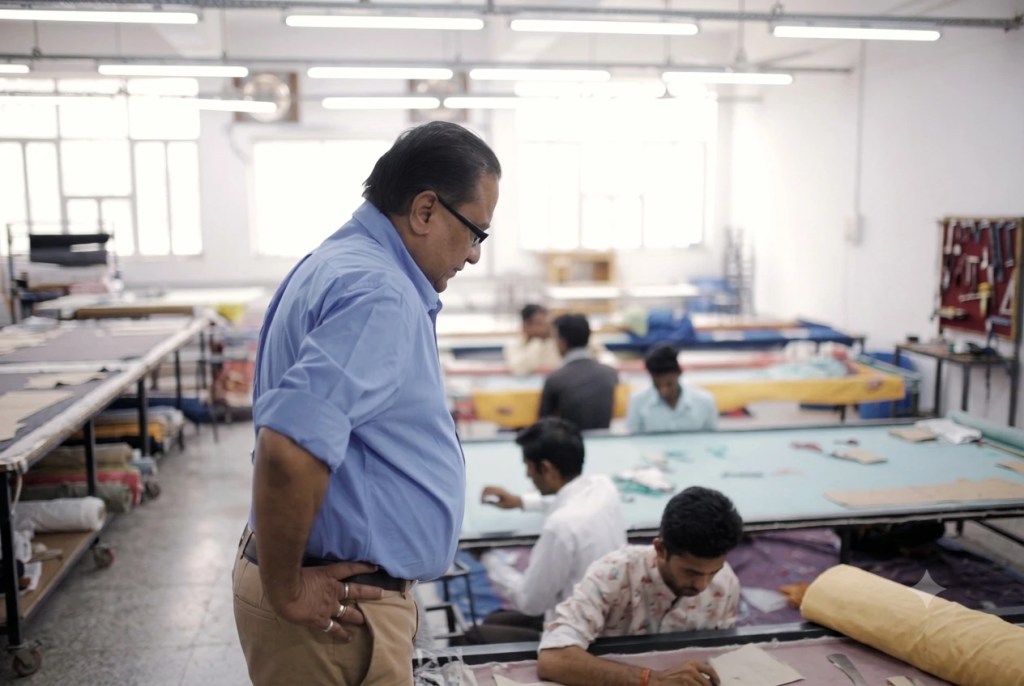 A man in a blue shirt and khaki pants stands in a workshop, looking down at a table where another man is working.