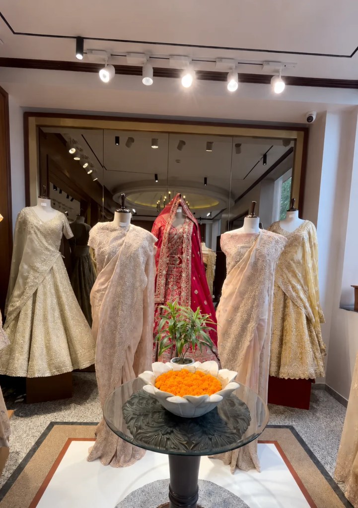 A store display featuring five mannequins showcasing traditional Indian clothing, with a table in the foreground holding a bowl of orange flowers.