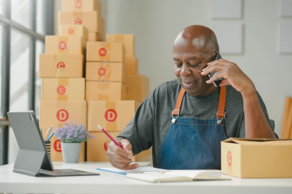 A man in a gray t-shirt and blue denim apron sits at a white desk, engaged in a phone call while writing with a red pencil.